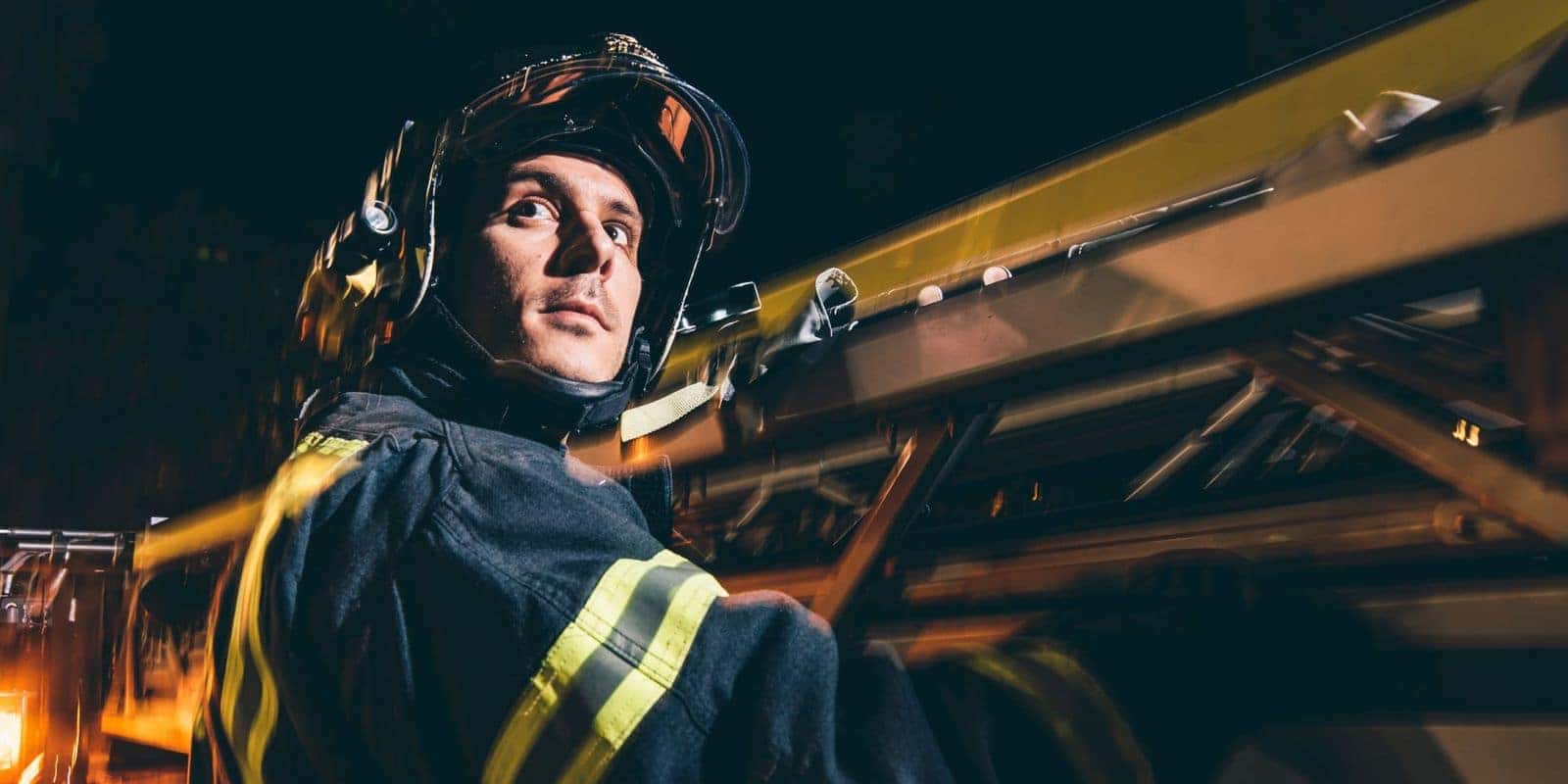 Firefighter in turnout gear and helmet on a fire truck ladder at night
