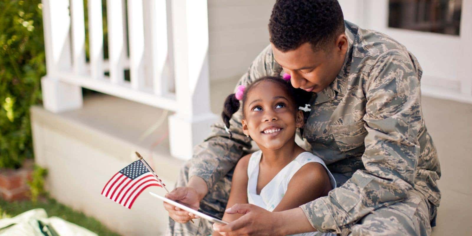 Service member in uniform sitting with his daughter, who holds a small American flag