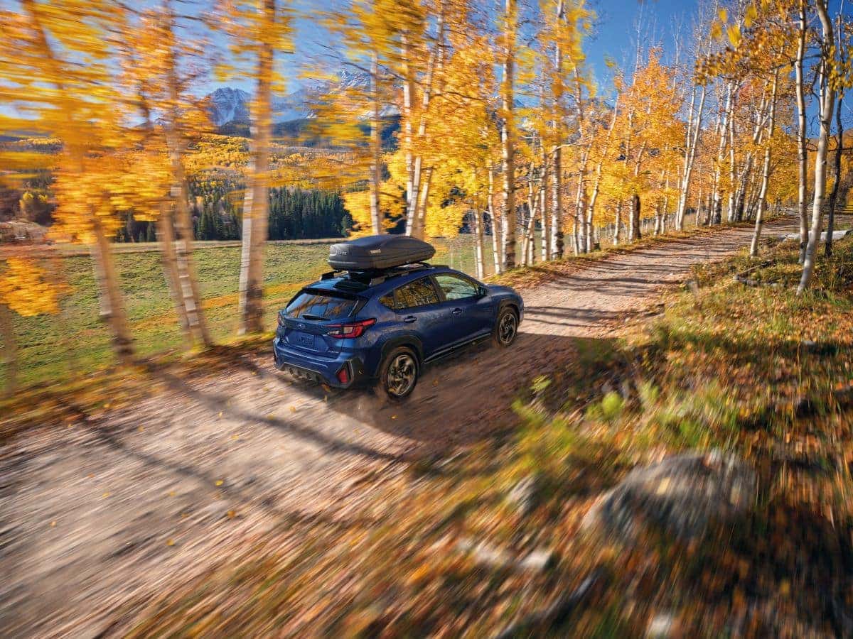 A dark blue Subaru Crosstrek with a rooftop cargo box drives along a dirt road lined with golden aspen trees and distant mountains in autumn