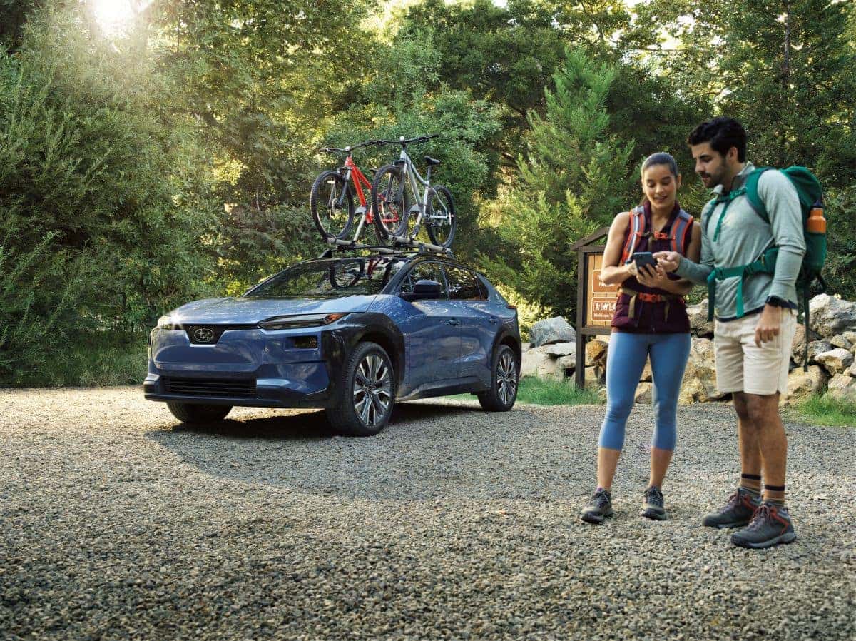 A couple in hiking gear checks a phone beside a blue Subaru Solterra with two bikes mounted on the roof rack at a wooded trailhead.