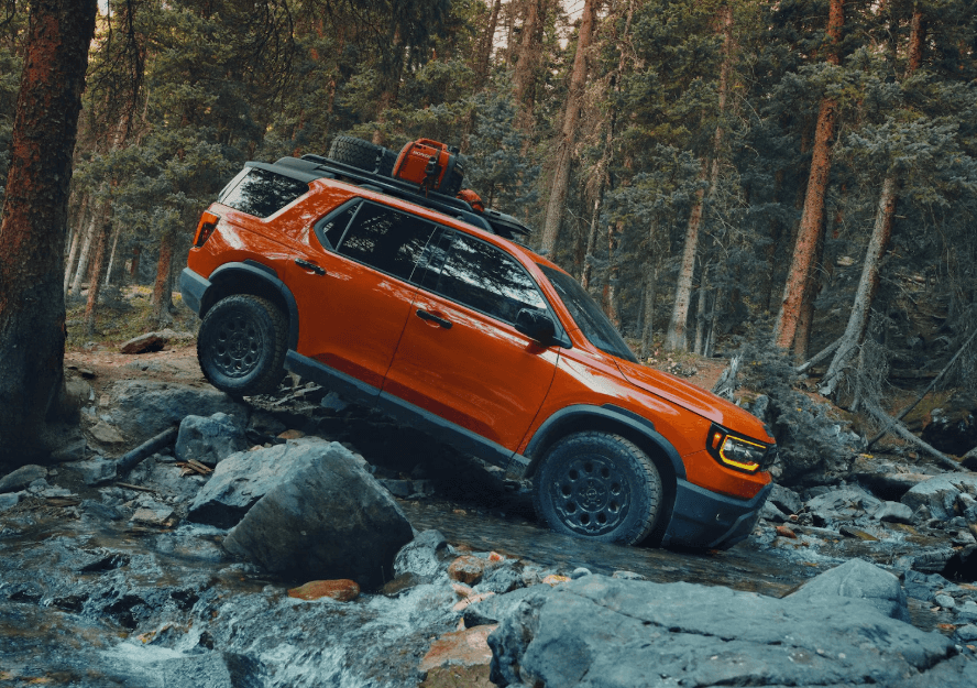 A vibrant orange 2026 Honda Passport TrailSport SUV climbs over rugged rocks while crossing a shallow creek in a dense forest, showcasing its off-road capability with all-terrain tires, roof rack, and mounted gear.