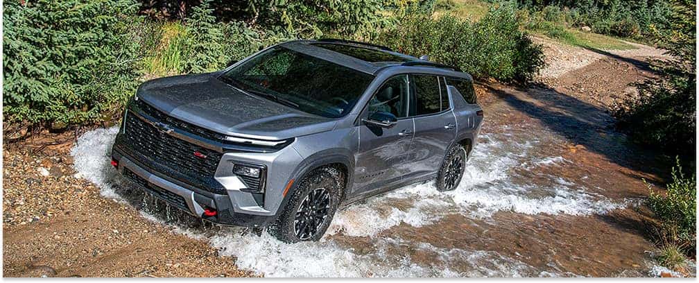 Front three-quarter view of a gray 2026 Chevrolet Traverse SUV driving through a shallow stream.