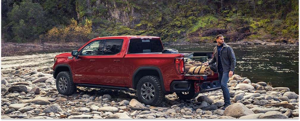Red 2026 GMC Sierra 1500 pickup truck parked on a rocky riverbank with the tailgate lowered.
