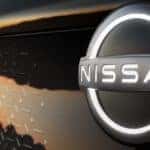A closeup shows the illuminated Nissan badging on the grille of a copper-colored Nissan Ariya at sunset.