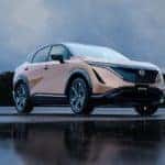 A pale orange 2021 Nissan Ariya is parked on a wet lot in front of trees near a Gallatin Nissan dealership.