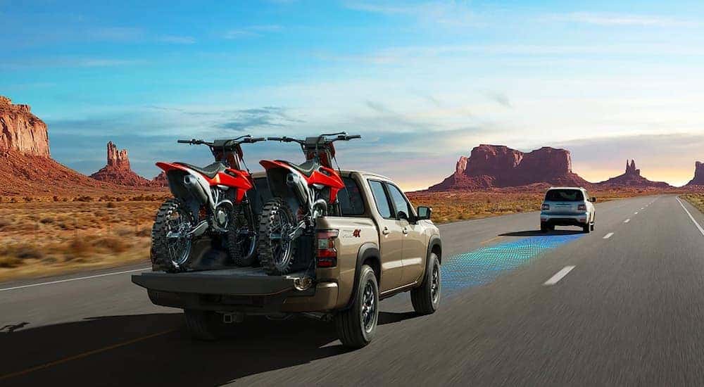 A tan 2022 Nissan Frontier Pro4x is shown on a highway with simulated sensor lines and dirt bikes in the bed.
