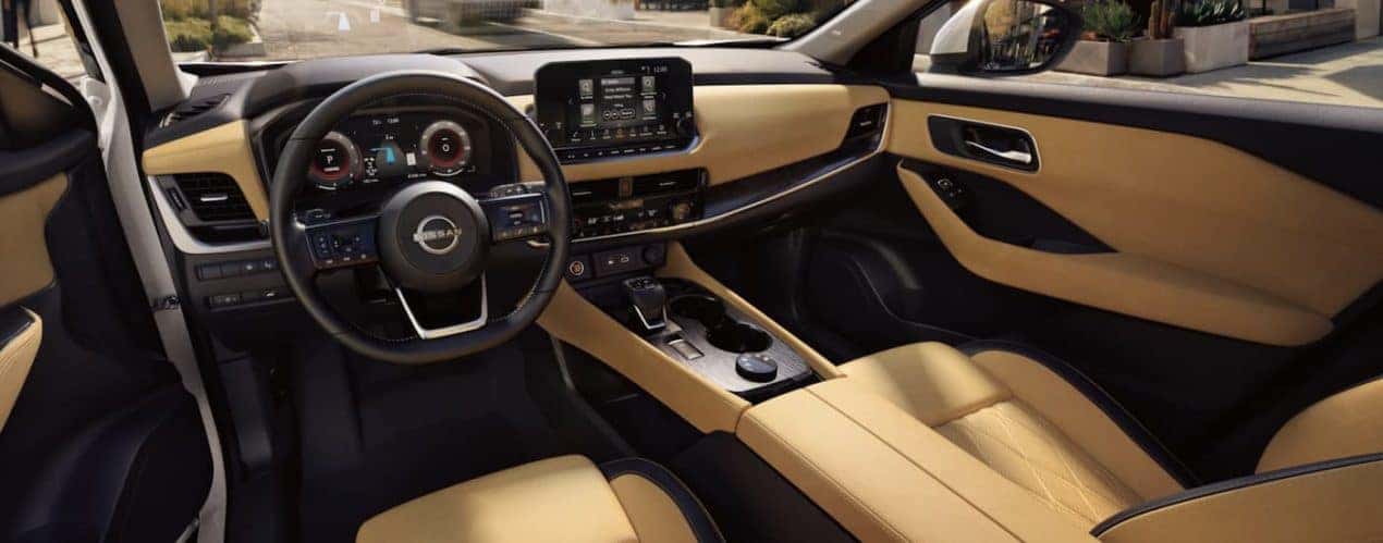 The black and tan interior of a 2022 Nissan Rogue shows the steering wheel and infotainment screen.