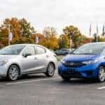 Two subcompact economy cars parked side by side on a sunlit suburban dealership lot, three-quarter front angle, one a silver hatchback-style sedan and one a small blue hatchback. Clean asphalt, light poles and autumn trees in the background under a bright overcast sky. Photorealistic automotive photography, even natural lighting, sharp detail on wheels and body lines. No text, no logos other than natural vehicle badging, no identifiable faces. (Representative image for illustration purposes only. Actual vehicle may vary based on trim and configuration.)