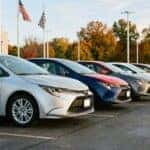 A row of new compact sedans in varied colors lined up neatly across a dealership front lot on a clear autumn morning, low three-quarter angle showing the nearest silver sedan in sharp focus with the row receding into soft focus. Clean pavement, flagpoles and trees with turning leaves in the background. Photorealistic automotive inventory photography, crisp golden-hour light. No text, no logos other than natural vehicle badging, no identifiable faces. (Representative image for illustration purposes only. Actual vehicle may vary based on trim and configuration.)
