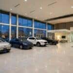 Photorealistic wide interior view of a bright modern car dealership showroom on a late-November evening: three new crossovers and a sedan arranged on polished pale tile under warm recessed lighting, tall glass frontage reflecting dark blue dusk and bare trees outside. Clean sales desks and a glass-walled delivery area in the background, floor freshly buffed, no shoppers present. Inviting retail-architecture photography, natural colour, wide-angle but undistorted. No text, no signage, no banners, no price stickers, no logos other than natural vehicle badging, no identifiable faces. (Representative image for illustration purposes only. Actual vehicle may vary based on trim and configuration.)