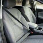 Photorealistic interior detail of a modern compact crossover SUV front seat: the driver's three-point seat belt drawn across a grey cloth seat, buckle latched into the receiver beside the centre console, the retractor pillar anchor visible at the B-pillar. Soft natural window light raking across the fabric, clean uncluttered cabin, shallow depth of field on the buckle. Calm, reassuring safety-documentation realism, natural colour. No people, no text, no signage, no logos other than natural vehicle badging, no identifiable faces. (Representative image for illustration purposes only. Actual vehicle may vary based on trim and configuration.)