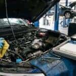 Photorealistic close-up inside a clean automotive service bay: an open engine bay of a modern sedan with the intake tubing detached, showing a throttle body and air filter housing being cleaned. A technician's gloved hands hold a microfibre cloth and an aerosol cleaner; a fresh cabin air filter, a new engine air filter and a brake fluid bottle sit on a parts tray beside the fender. Crisp daylight mixed with overhead shop lighting, shallow depth of field. Documentary maintenance realism, natural colour. No text, no signage, no logos other than natural vehicle badging, no identifiable faces. (Representative image for illustration purposes only. Actual vehicle may vary based on trim and configuration.)