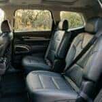 Photorealistic second-row interior of a modern three-row family SUV photographed through the open rear door: two outboard seat belts hanging from their B- and C-pillar anchors across dark leather captain's chairs, lower anchor points and buckle stalks clearly visible between the seats. Warm late-afternoon light entering through the open door, immaculate empty cabin, third row folded flat behind. Careful, factual safety-inspection realism, natural colour, moderate depth of field. No people, no text, no signage, no logos other than natural vehicle badging, no identifiable faces. (Representative image for illustration purposes only. Actual vehicle may vary based on trim and configuration.)