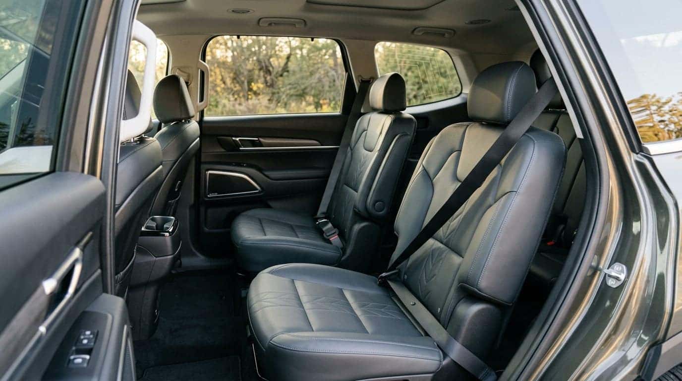 Photorealistic second-row interior of a modern three-row family SUV photographed through the open rear door: two outboard seat belts hanging from their B- and C-pillar anchors across dark leather captain's chairs, lower anchor points and buckle stalks clearly visible between the seats. Warm late-afternoon light entering through the open door, immaculate empty cabin, third row folded flat behind. Careful, factual safety-inspection realism, natural colour, moderate depth of field. No people, no text, no signage, no logos other than natural vehicle badging, no identifiable faces. (Representative image for illustration purposes only. Actual vehicle may vary based on trim and configuration.)