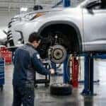 Photorealistic dealership service bay: a silver midsize three-row SUV raised on a hydraulic lift with its front wheel removed, revealing the front brake caliper and rotor. A certified technician in clean workwear and safety glasses, seen from behind and to the side, inspects the caliper with a torque wrench. Bright overhead shop lighting, polished concrete floor, tidy tool chest in the soft-focus background. Documentary service-department realism, natural colour, shallow depth of field. No text, no signage, no logos other than natural vehicle badging, no identifiable faces. (Representative image for illustration purposes only. Actual vehicle may vary based on trim and configuration.)