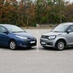Photorealistic three-quarter front view of two small vehicles parked side by side on a clean paved lot: on the left a compact four-door economy sedan in deep blue, on the right a boxy subcompact crossover in light grey with roof rails. Both at matching angles so their body styles contrast clearly, sedan roofline low and crossover upright. Overcast soft daylight, low autumn treeline and plain asphalt behind, no bystanders. Clean automotive-comparison photography, natural colour, even exposure. No text, no signage, no logos other than natural vehicle badging, no identifiable faces. (Representative image for illustration purposes only. Actual vehicle may vary based on trim and configuration.)