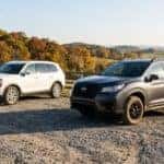 Photorealistic outdoor view of two midsize three-row family SUVs photographed from a low front three-quarter angle, parked a few feet apart on a gravel pull-off at the edge of rolling Tennessee farmland: the nearer one in dark grey with a rugged off-road-style stance and raised ride height, the farther one in white with a smoother suburban profile. Early morning light, long shadows, split-rail fence and autumn hardwoods behind. Honest comparison-review photography, natural colour, both vehicles sharply lit and fully visible. No text, no signage, no logos other than natural vehicle badging, no identifiable faces. (Representative image for illustration purposes only. Actual vehicle may vary based on trim and configuration.)