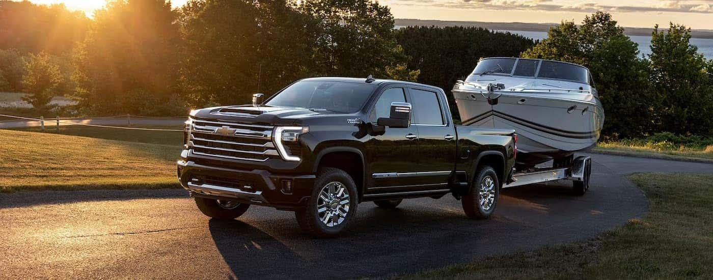 A black 2024 Chevy Silverado 2500HD High Country is shown towing a white boat on a winding road.