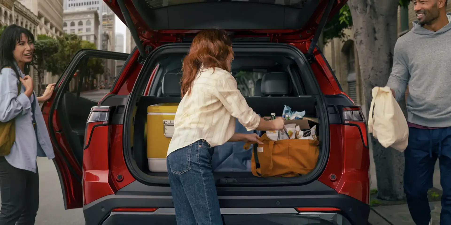 Friends loading a cooler and bags into the back of a red Chevrolet SUV on a city street