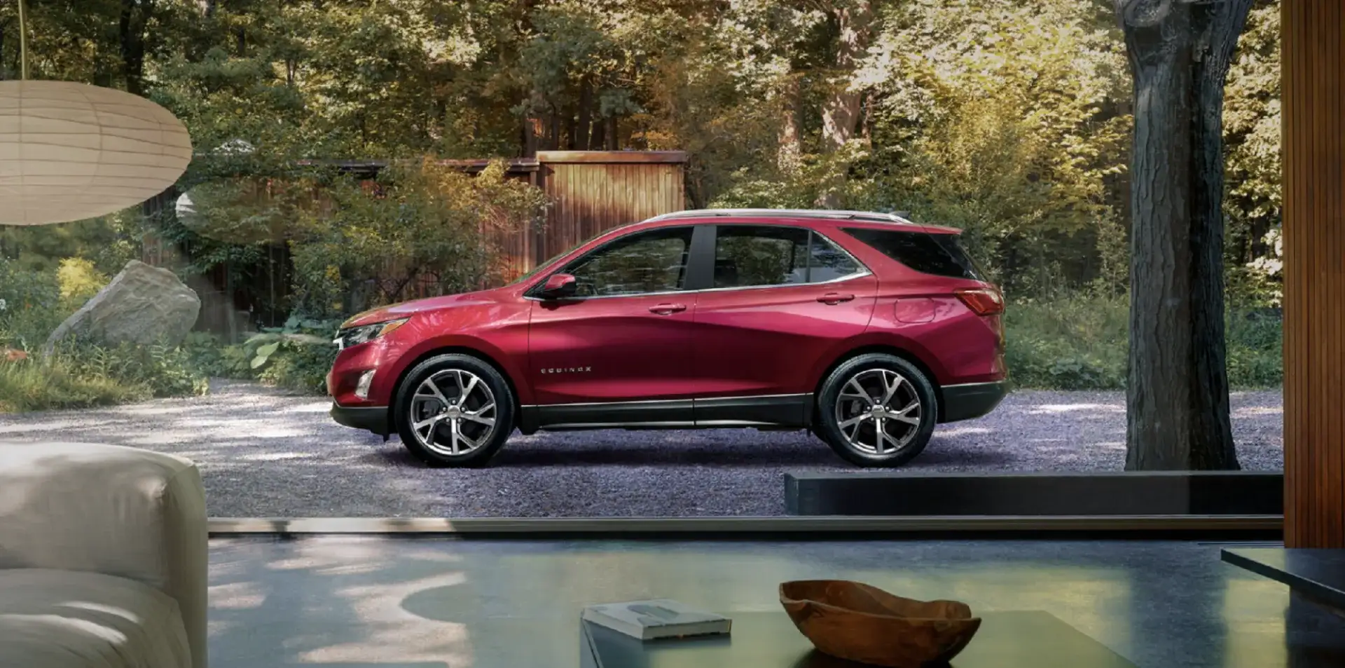 Chevrolet Equinox parked on a gravel driveway surrounded by trees, viewed through a modern living room window